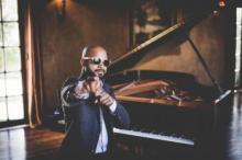 Barron Ryan seated at grand piano with sunglasses on pointing at camera